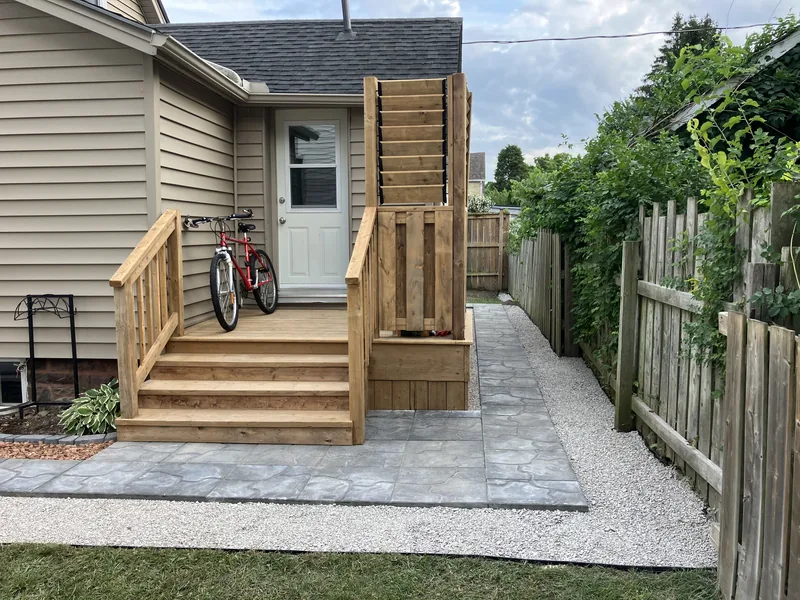 After: new wood landing and stairs at the side door, with railings, a slatted privacy screen, and a paver walkway along the fence