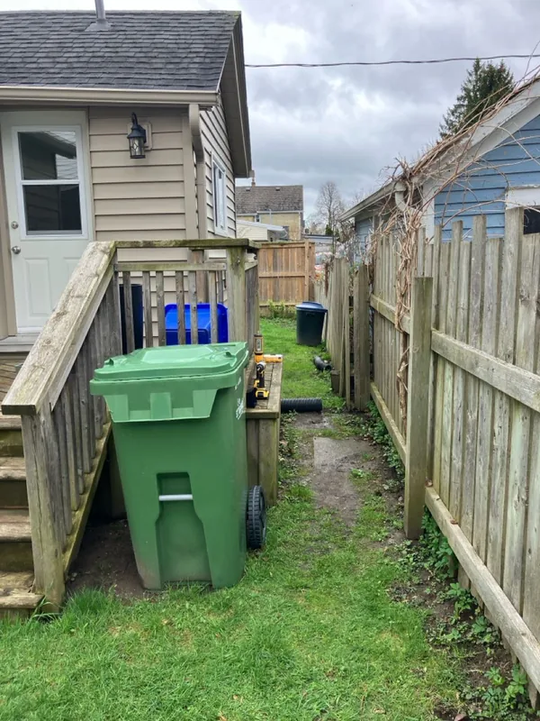 Before: old grey stairs at the side door, with garbage and recycling bins against the railing and a muddy path along the fence